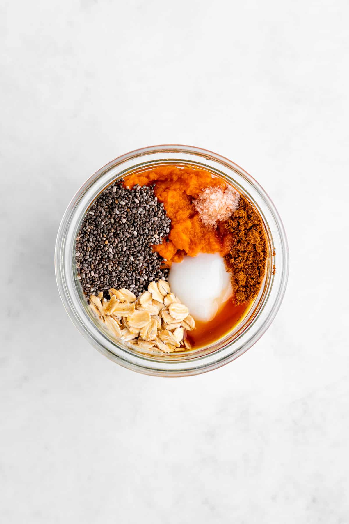 overhead photo of a mason jar filled with rolled oats, pumpkin puree, yogurt, chia seeds, cinnamon, and maple syrup