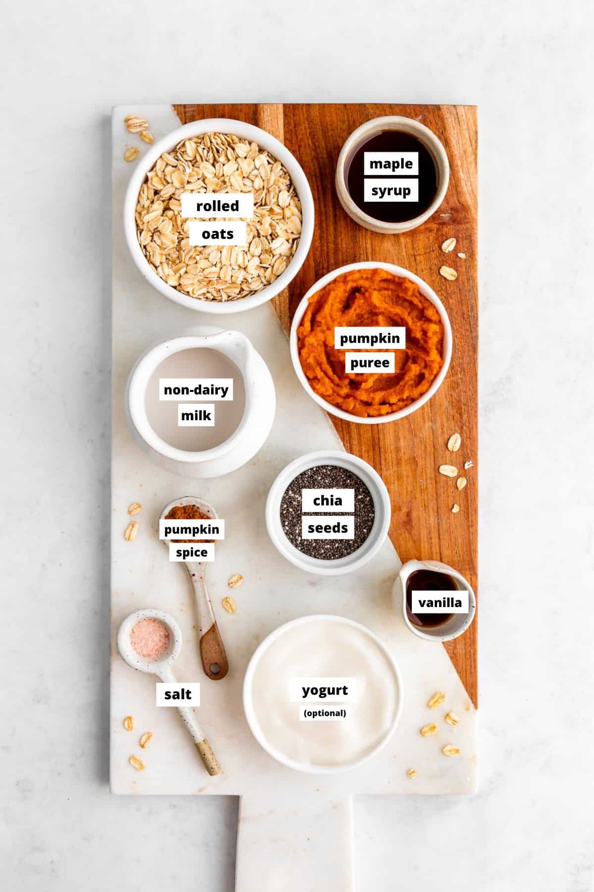 a marble board topped with bowls of oats, pumpkin puree, yogurt, chia seeds, pumpkin spice, maple syrup, and almond milk
