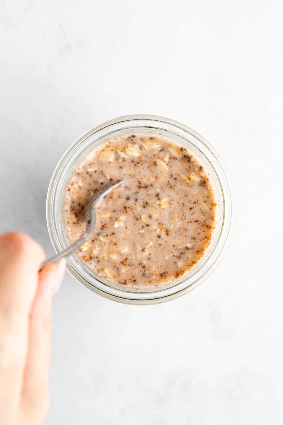 left hand holding a spoon mixing vegan vanilla overnight oats in a jar