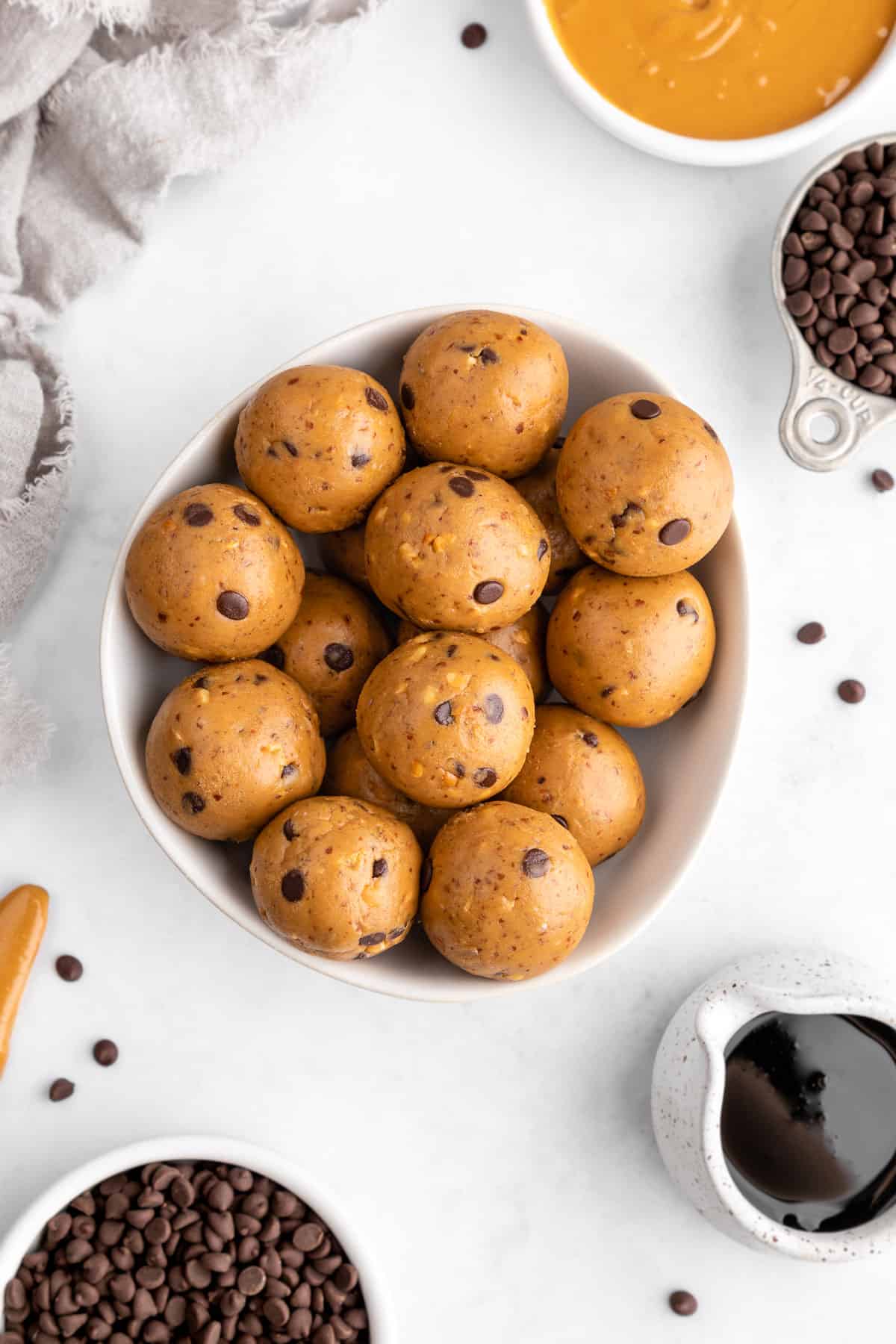 a bowl of peanut butter protein balls surrounded by chocolate chips, peanut butter, and maple syrup