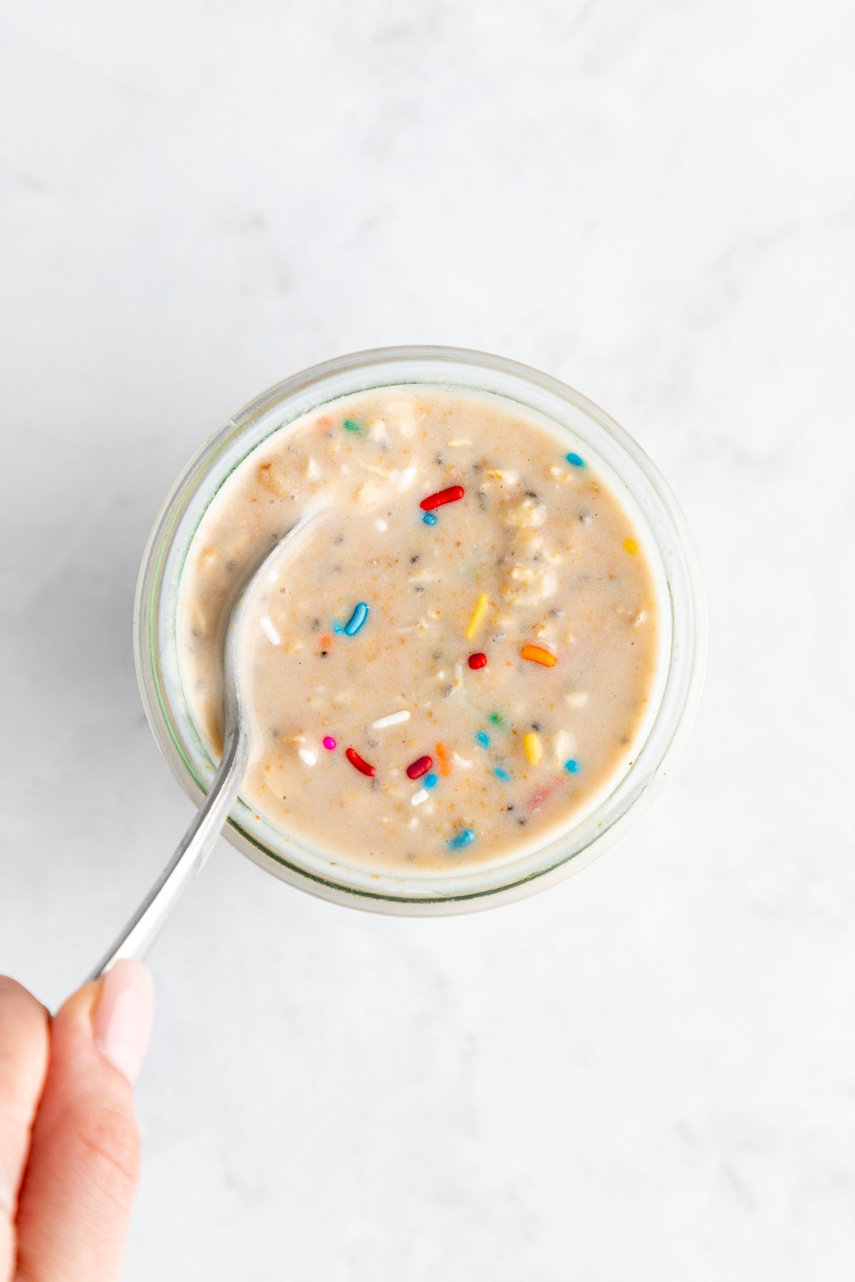 a hand holding a spoon mixing birthday cake overnight oats in a jar