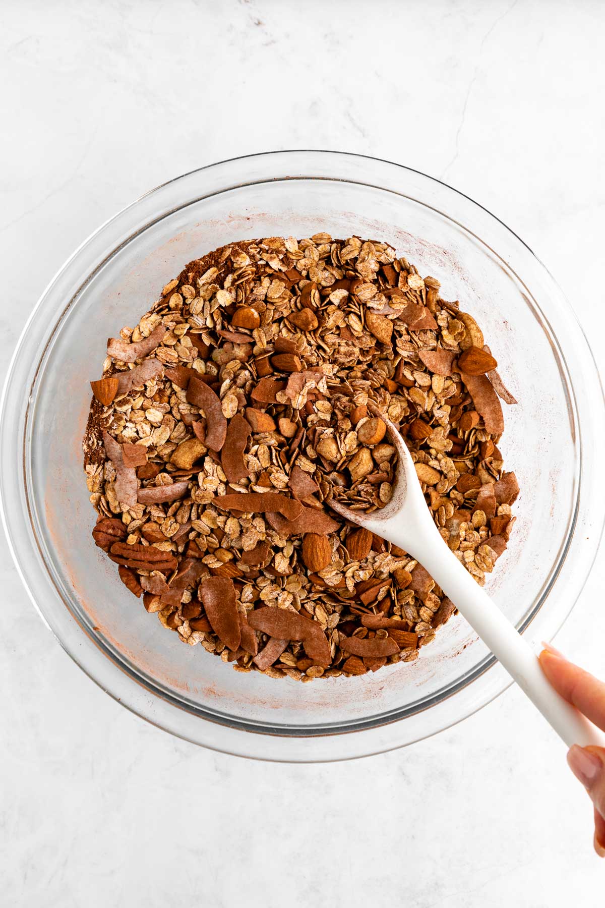 hand holding a wooden spoon mixing rolled oats, almonds, coconut, cocoa powder, and coconut sugar in a bowl