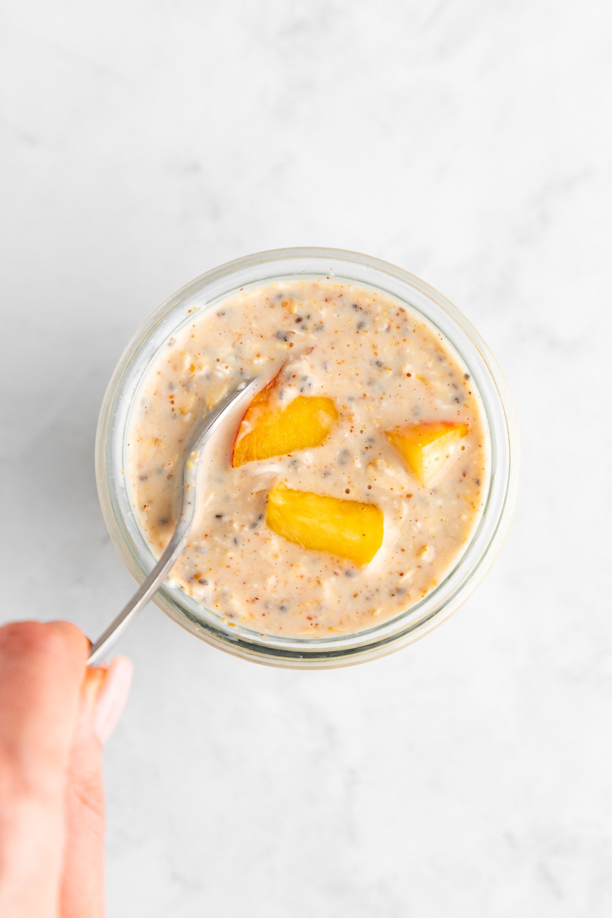 a hand holding a spoon mixing peaches and cream overnight oats in a jar