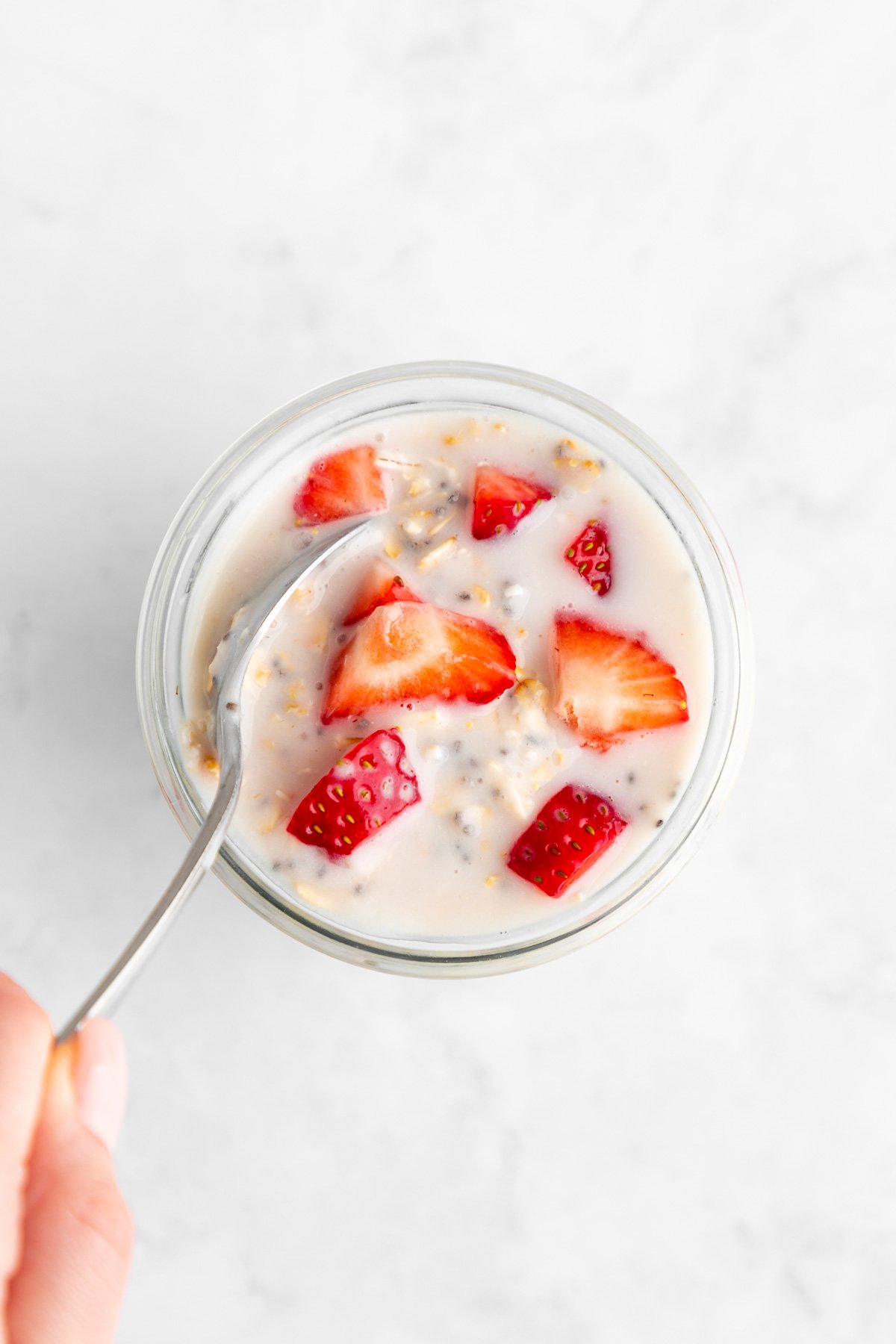 a hand holding a spoon mixing strawberries and cream overnight oats in a jar