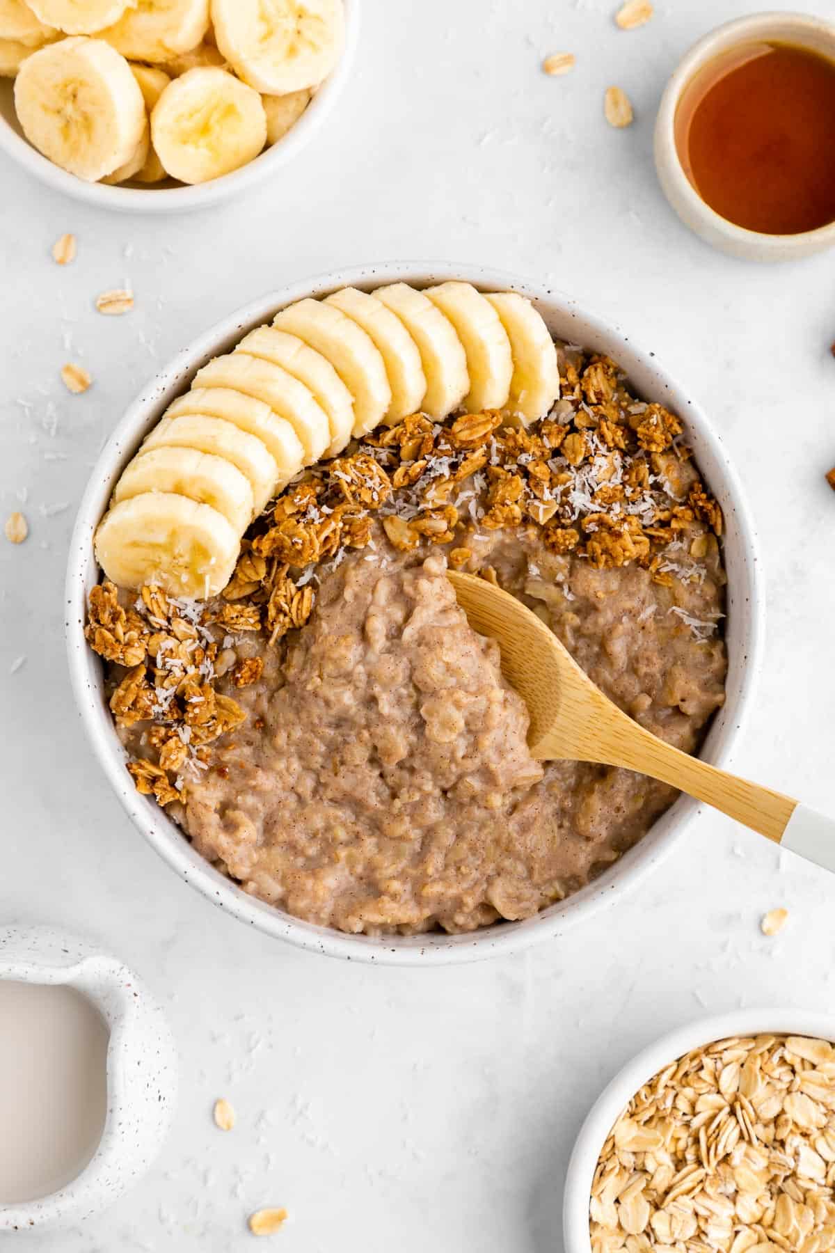 a wooden spoon scooping into a bowl of vegan banana oatmeal surrounded by bowls of rolled oats, sliced banana, almond milk, and maple syrup