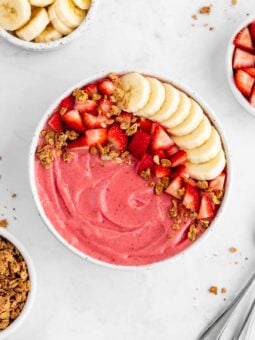 a strawberry banana smoothie bowl surrounded by almond milk, granola, and fresh fruit