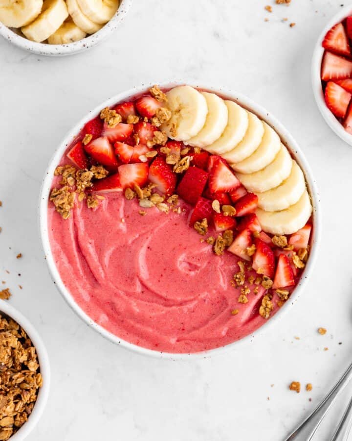 a strawberry banana smoothie bowl surrounded by almond milk, granola, and fresh fruit