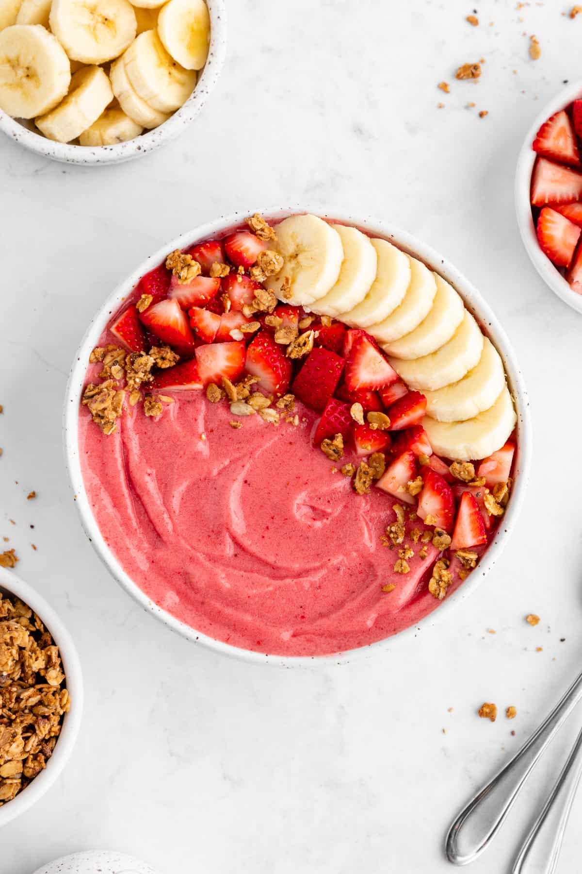 a strawberry banana smoothie bowl surrounded by almond milk, granola, and fresh fruit