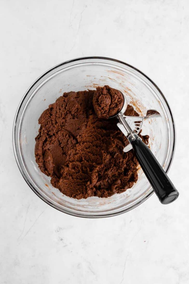 a cookie scoop scooping vegan chocolate cookie dough in a glass bowl