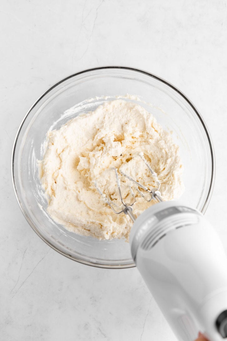 a handheld electric mixer creaming vegan butter, vegan cream cheese, and vanilla extract together