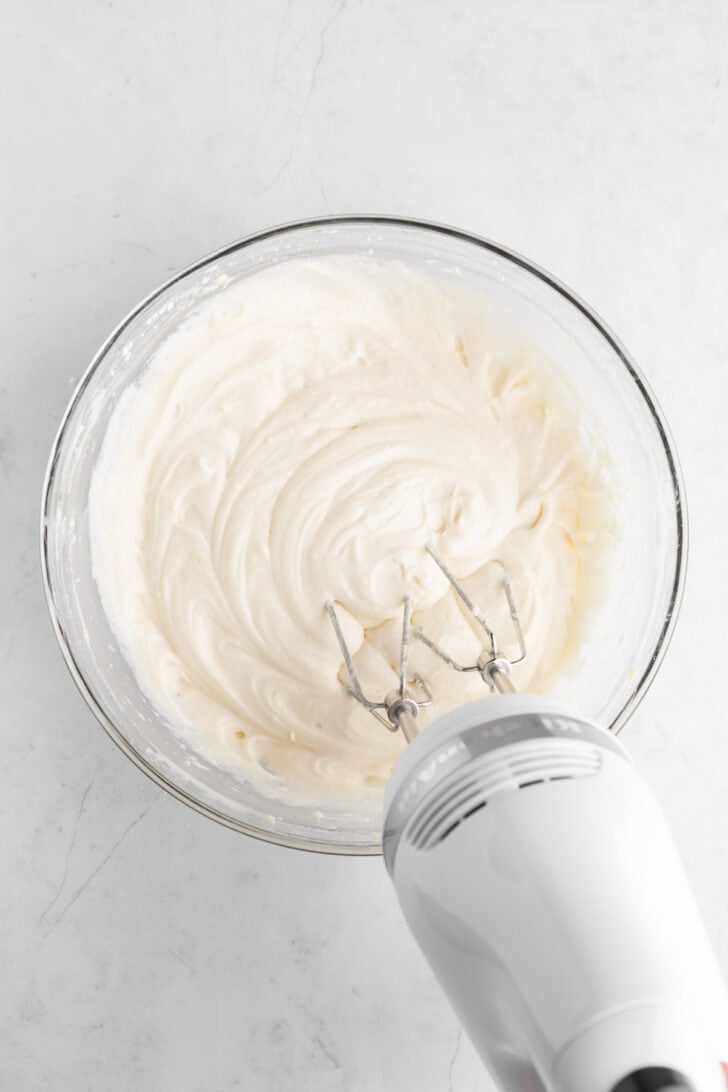 a handheld electric mixer beating homemade vegan cream cheese frosting in a glass mixing bowl