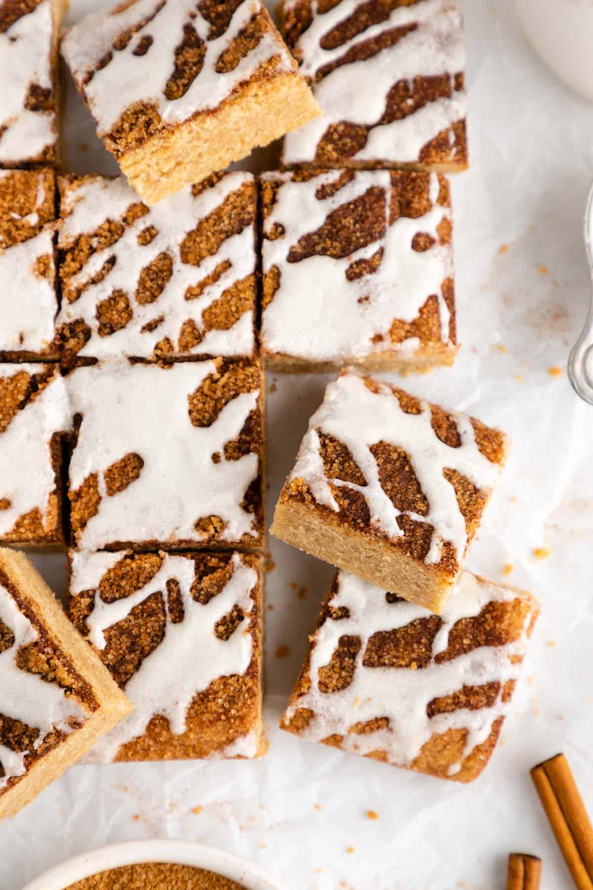 a pile of vegan cinnamon roll blondies with cinnamon sugar and vanilla icing on white parchment paper