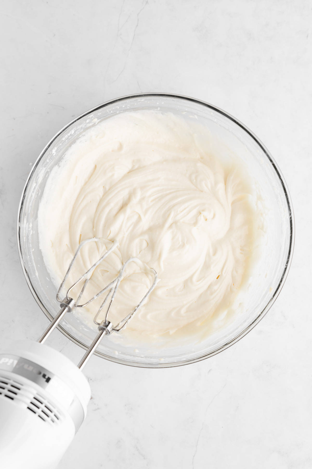 a handheld electric mixer beating homemade vegan cream cheese frosting in a glass mixing bowl