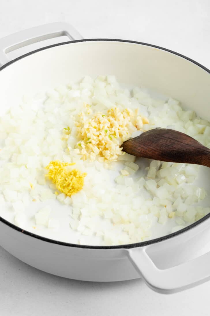 a wooden spoon sautéing diced onion, garlic, and ginger in a white le creuset dutch oven