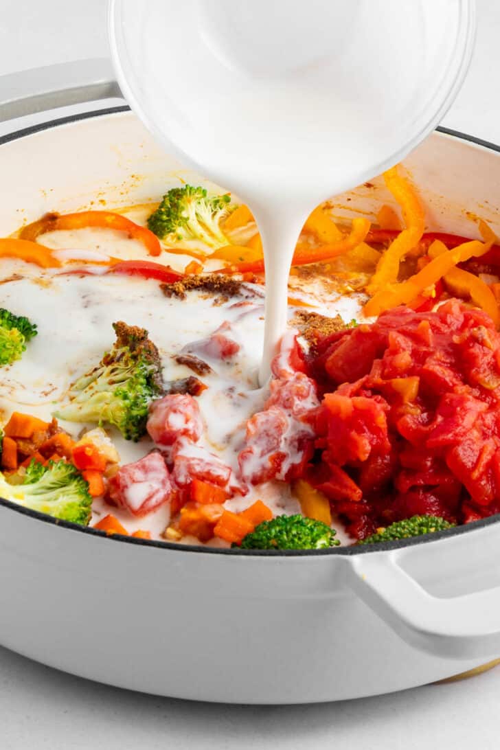 pouring coconut milk over vegetables in a white le creuset dutch oven