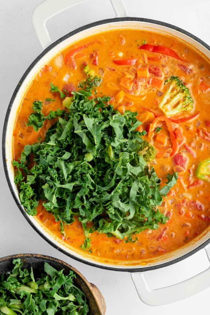 leafy kale added to vegan thai red curry cooking in a white le creuset dutch oven