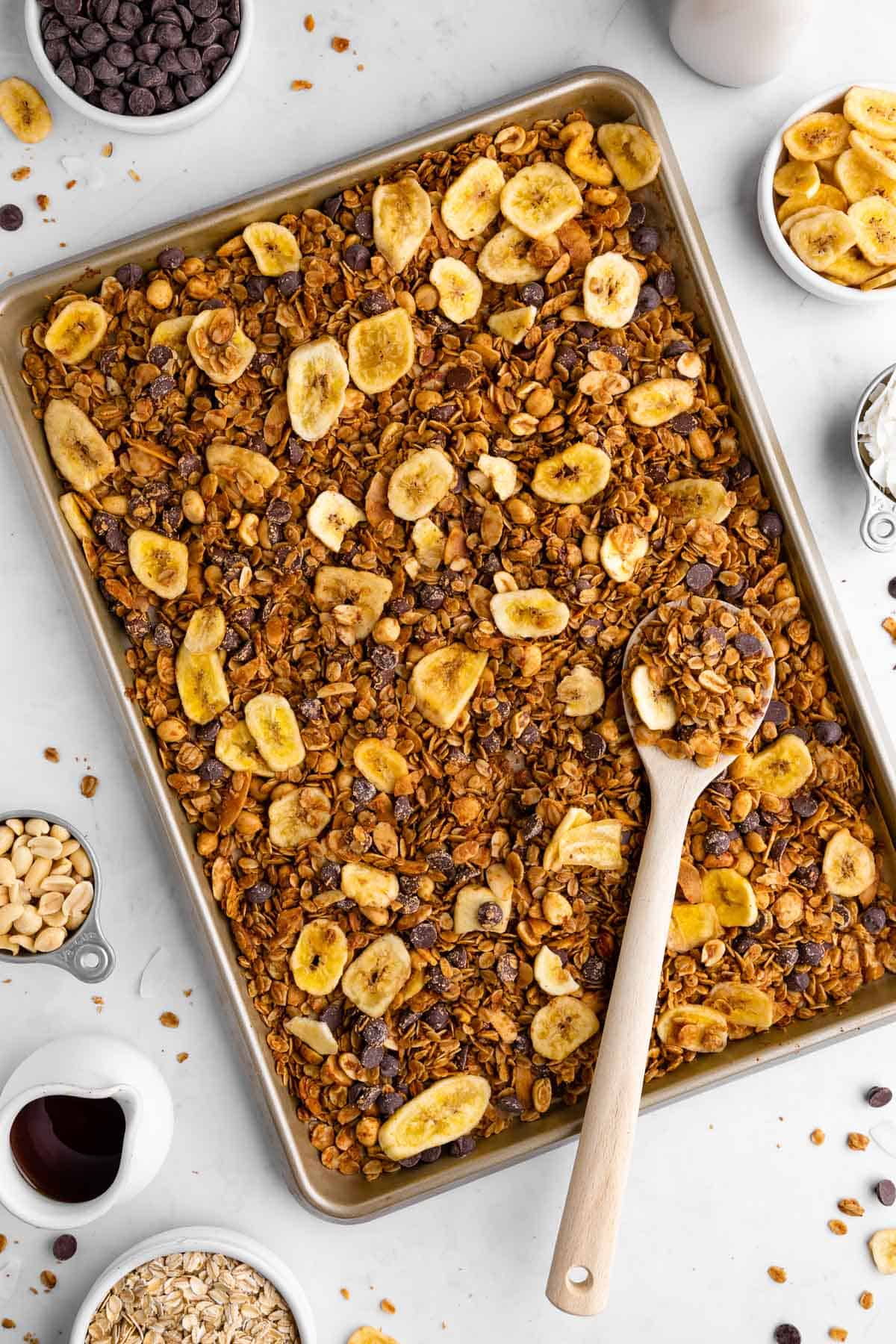 homemade chunky monkey granola with banana chips, nuts, coconut, and chocolate chips on a baking sheet