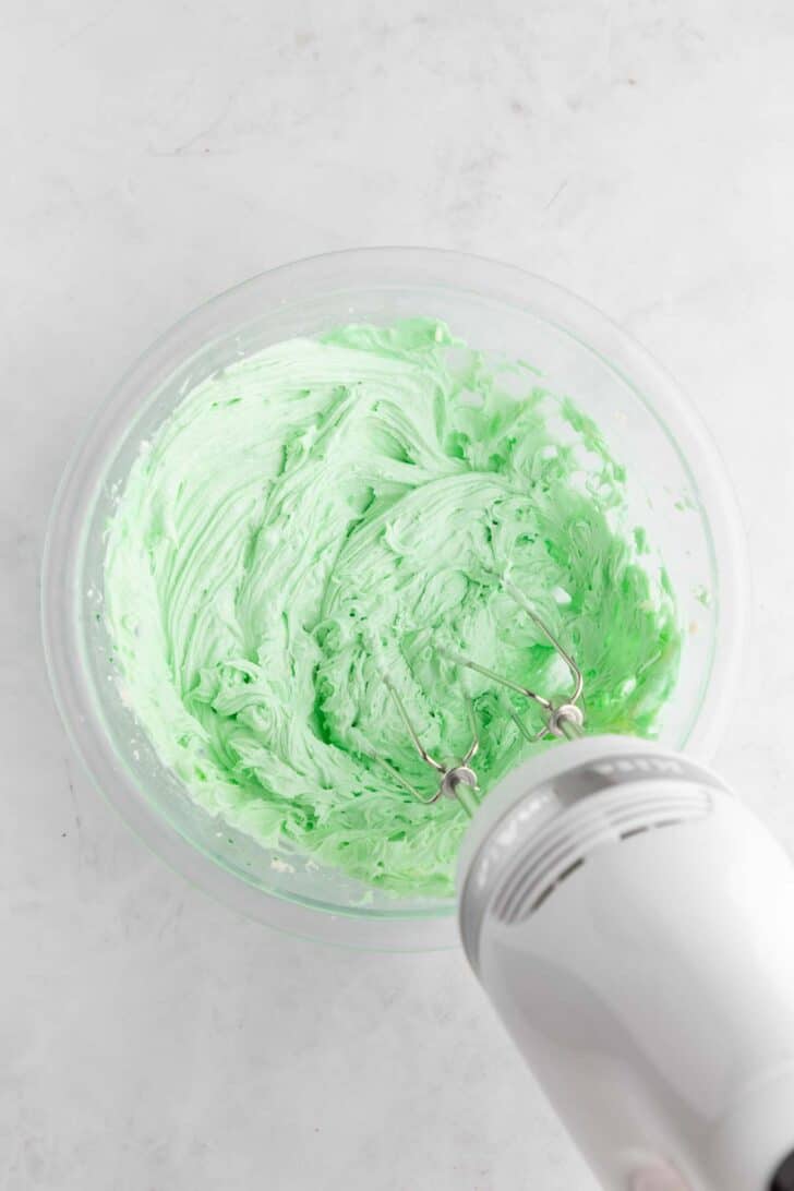 a kitchenaid handheld mixer creaming mint buttercream frosting in a glass bowl