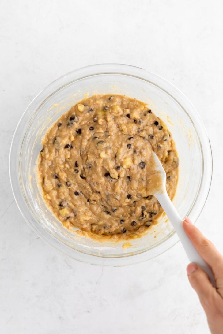 mixing vegan banana bread batter with chocolate chips and walnuts inside a glass bowl