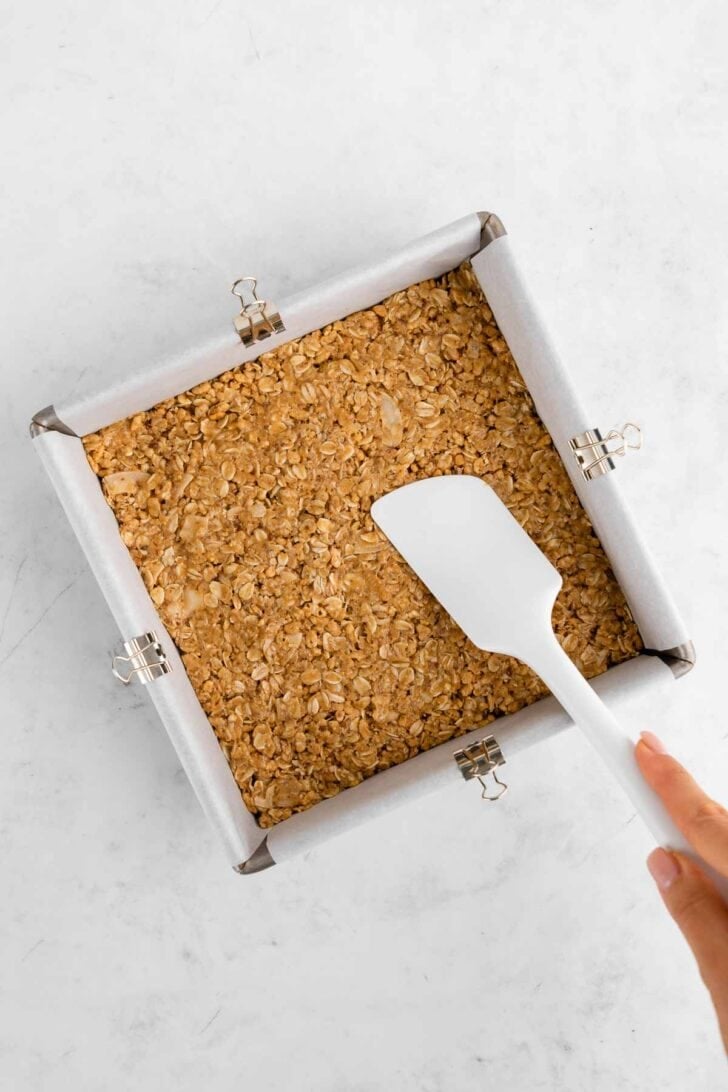 pressing homemade coconut chocolate chip granola bars into a baking pan with a rubber spatula